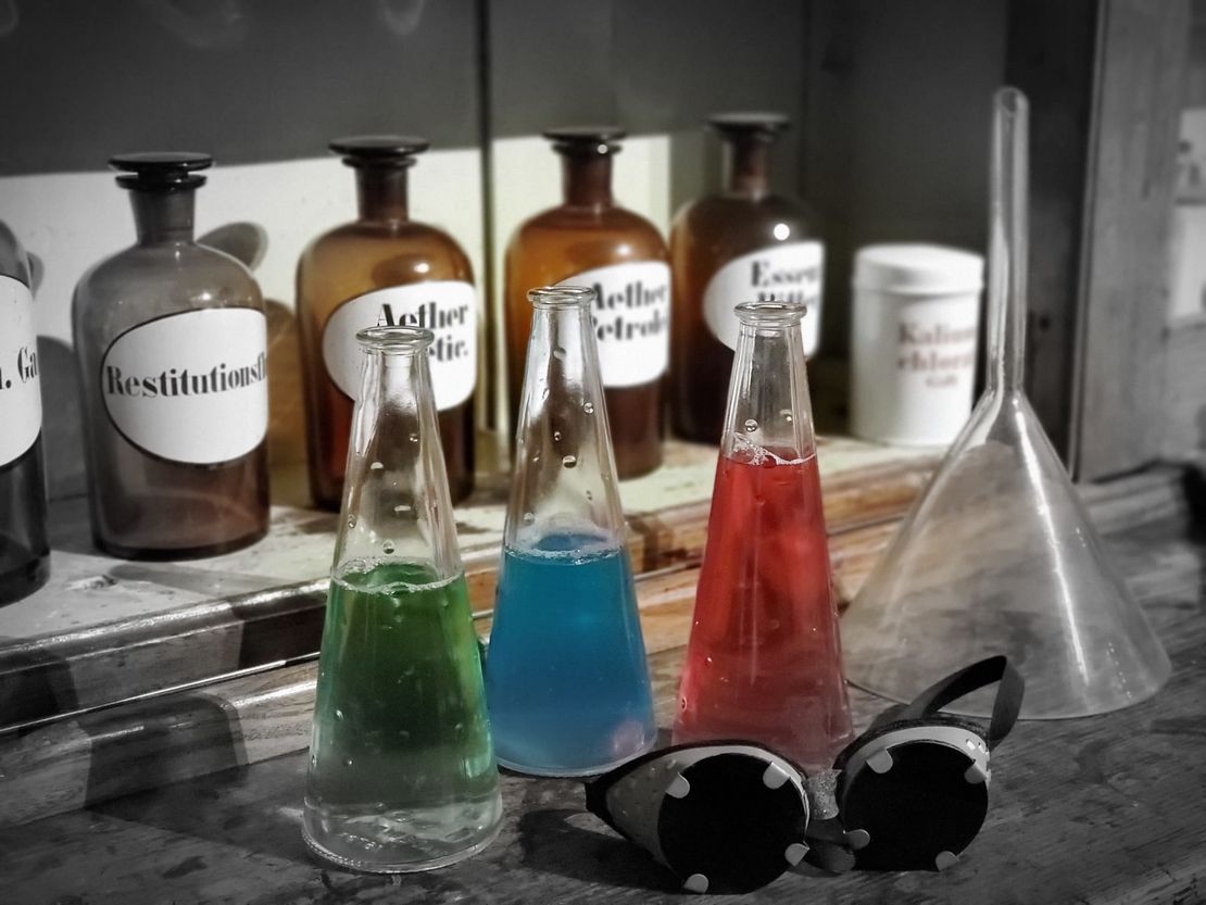 Drei Flaschen mit farbiger Flüssigkeit, eine Brille mit dunklem Glas, ein Glastrichter und im Hintergrund alte Apothekerflaschen.