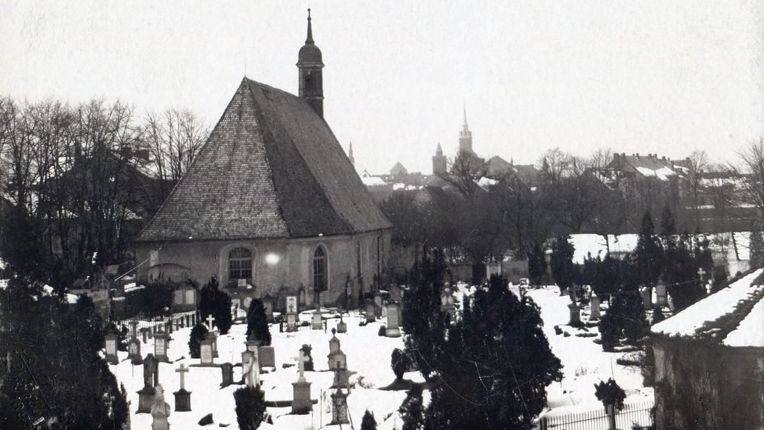 Schwarzweißfoto einer kleinen Kirche. Davor Gräber, die mit Schnee bedeckt sind.
