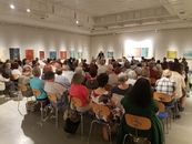 Viele Leute sitzen in einem großen Saal im Museum und hören einer Ansprache zu. Ringsum sieht man bunte Bilder an den Wänden.