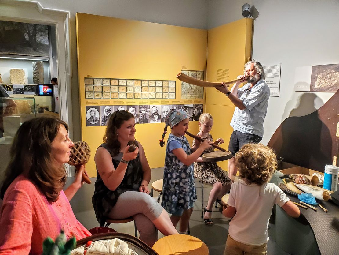 Mütter und Kleinkinder mit diversen Musikinstrumenten aus natürlichen Materialien wie Horn oder Holz und Museumspädagoge Ulrich Schollmeyer mit einem großen Horn.