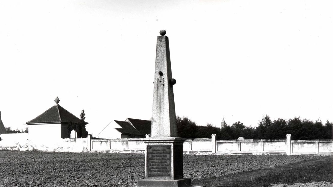 Schwarzweißfoto eines obeliskartigen Kriegerdenkmals aus Stein auf einem Sockel mit Inschrift. Das Denkmal steht auf einem Feld, dahinter ist die Friedhofsmauer zu sehen.