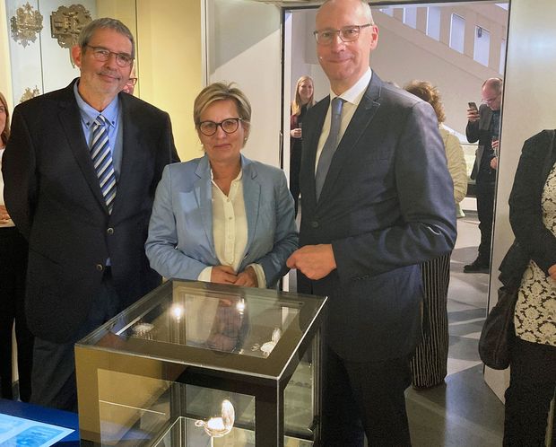 Ein Mann, eine Frau und noch ein Mann stehen in dunkelblauen Anzügen gekleidet vor einer Glaskiste. Sie lächeln in die Kamera