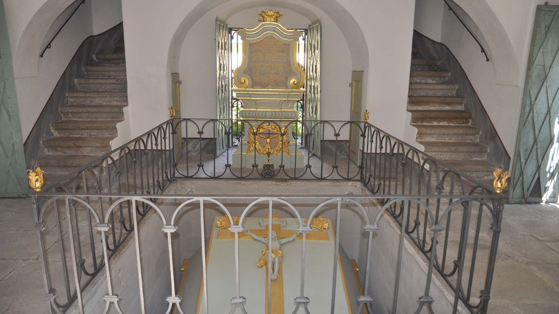 Eingangsbereich eines alten Gebäudes. Links und rechts gehen Treppen in das erste Stockwerk. Im Boden ist steinerner Sarkophag mit einem goldenen Kreuz eingelassen und rundum von einem Geländer umgeben.